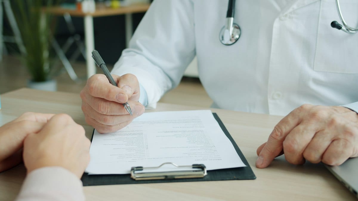 Un médecin en blouse blanche, stéthoscope autour du cou, remplit un formulaire sur un presse-papiers face à un patient lors d'une consultation.