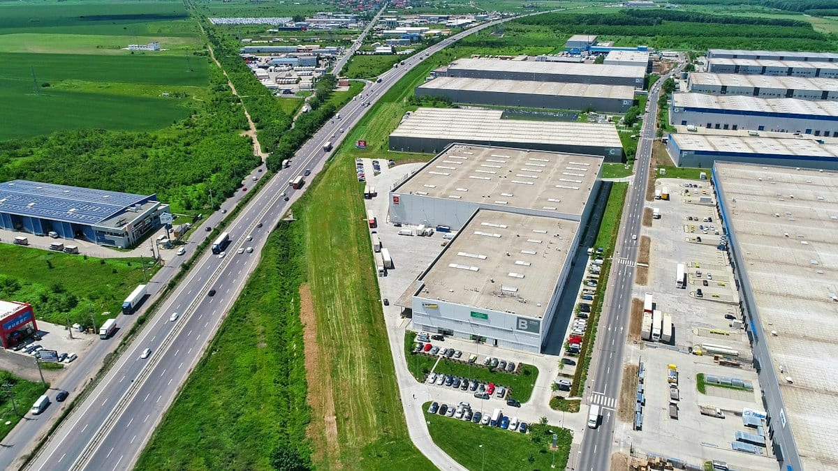 Vue aérienne d'une zone d'activités avec plusieurs entrepôts, des parkings et une route à grande circulation bordée de champs.