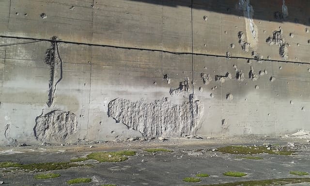 Mur de béton de la base sous-marine de Bordeaux criblé d'impacts et d'éclats laissés par les bombardements, au-dessus d'un sol envahi de mousse.