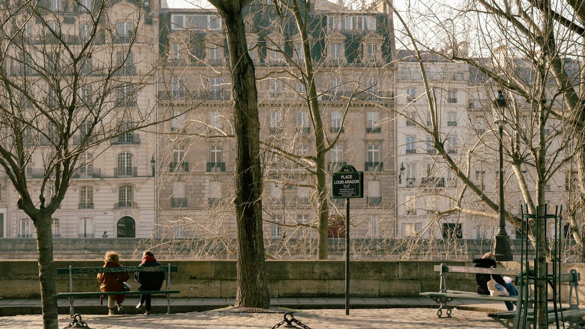 Place bordée d'arbres et de bancs où des personnes sont assises, devant des immeubles de pierre, illustrant un espace public ombragé et convivial.