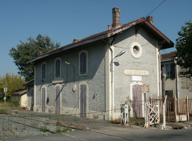 Façade de l'ancienne gare de la Souys à Floirac, bâtiment en crépi gris à volets violets, avec l'inscription « La Souys » et les anciens rails au premier plan.