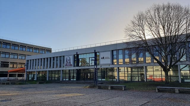 Façade vitrée d'un bâtiment du campus de l'université Bordeaux Montaigne à Pessac, avec son parvis dallé et un arbre nu au premier plan