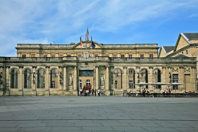 Façade de l'hôtel de ville de Bordeaux, place Pey-Berland, avec ses drapeaux et quelques passants devant l'entrée.