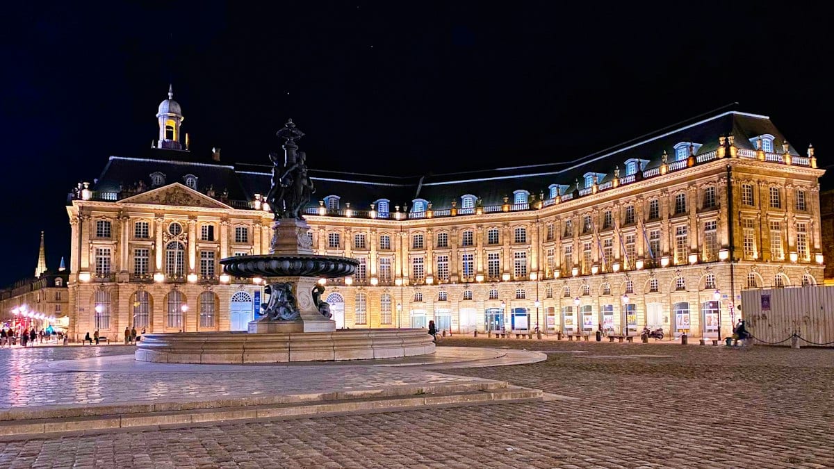 La place de la Bourse à Bordeaux de nuit, avec sa façade illuminée et la fontaine des Trois Grâces au premier plan.