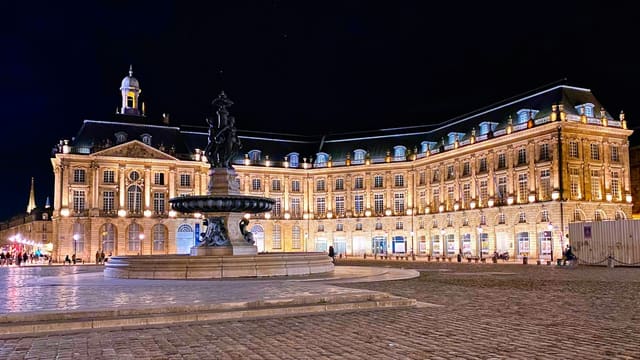 La place de la Bourse à Bordeaux de nuit, avec sa façade illuminée et la fontaine des Trois Grâces au premier plan.