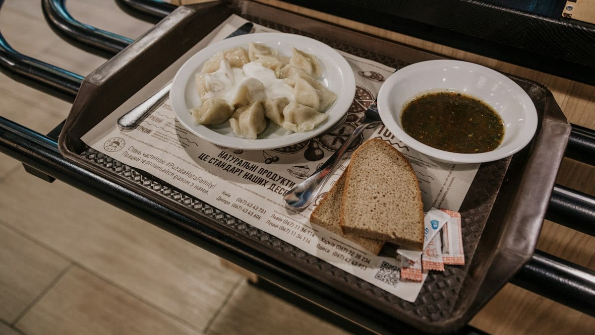 Plateau de cantine posé sur un rail en self-service, avec une assiette de raviolis, un bol de soupe et deux tranches de pain.