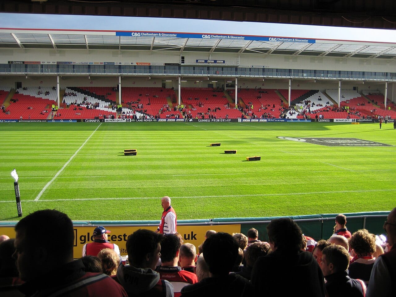 Tribunes du Kingsholm Stadium de Gloucester, pelouse rayée et supporters au premier plan avant un match de rugby.