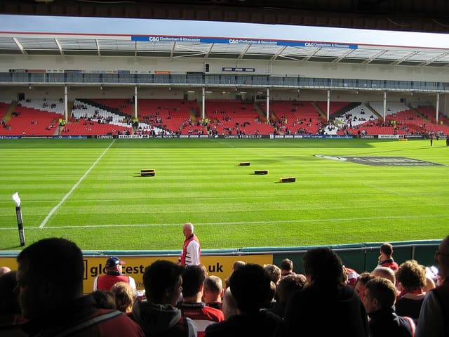 Tribunes du Kingsholm Stadium de Gloucester, pelouse rayée et supporters au premier plan avant un match de rugby.