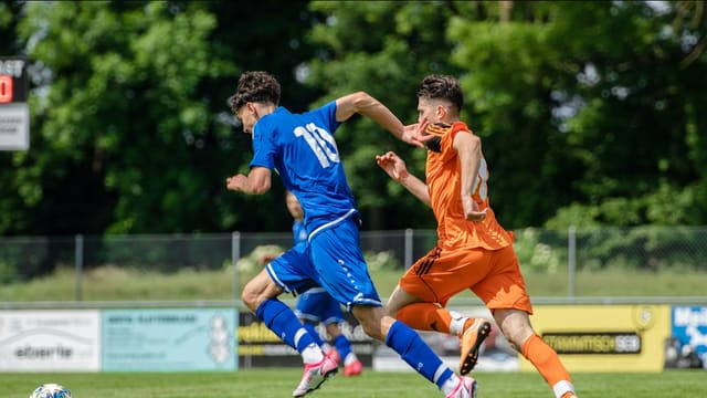 Deux joueurs en maillot bleu et orange se disputent le ballon lors d'un match de football amateur sur un terrain bordé d'arbres.