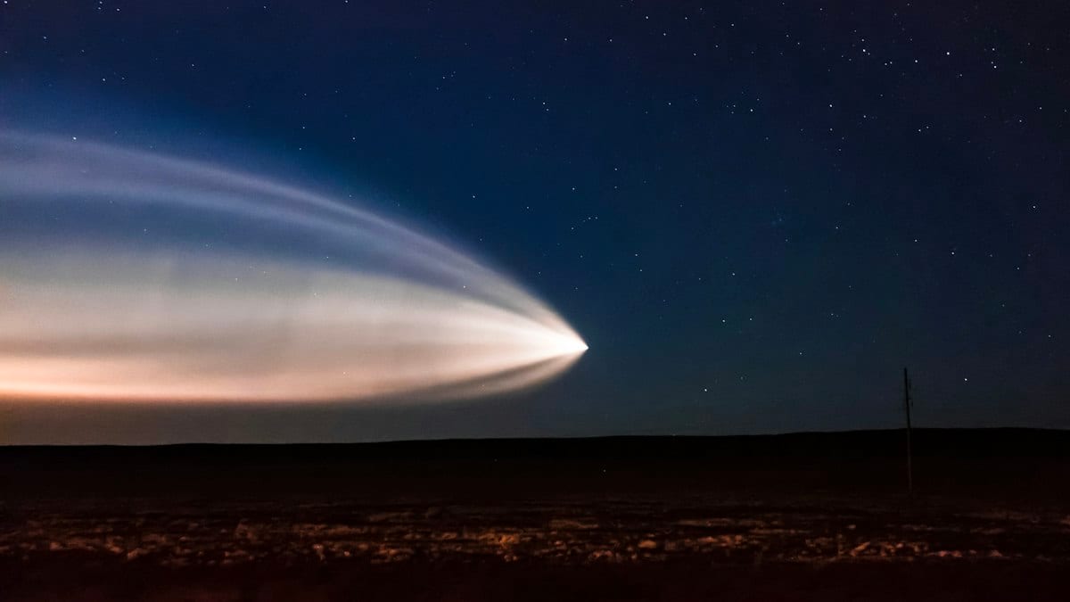 Longue traînée lumineuse d'un lancement de fusée dans un ciel étoilé, au-dessus d'un paysage sombre.