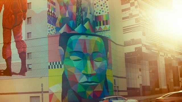 Grande fresque colorée sur la façade d'un immeuble, avec un visage géométrique aux yeux clos et un personnage en combinaison orange.
