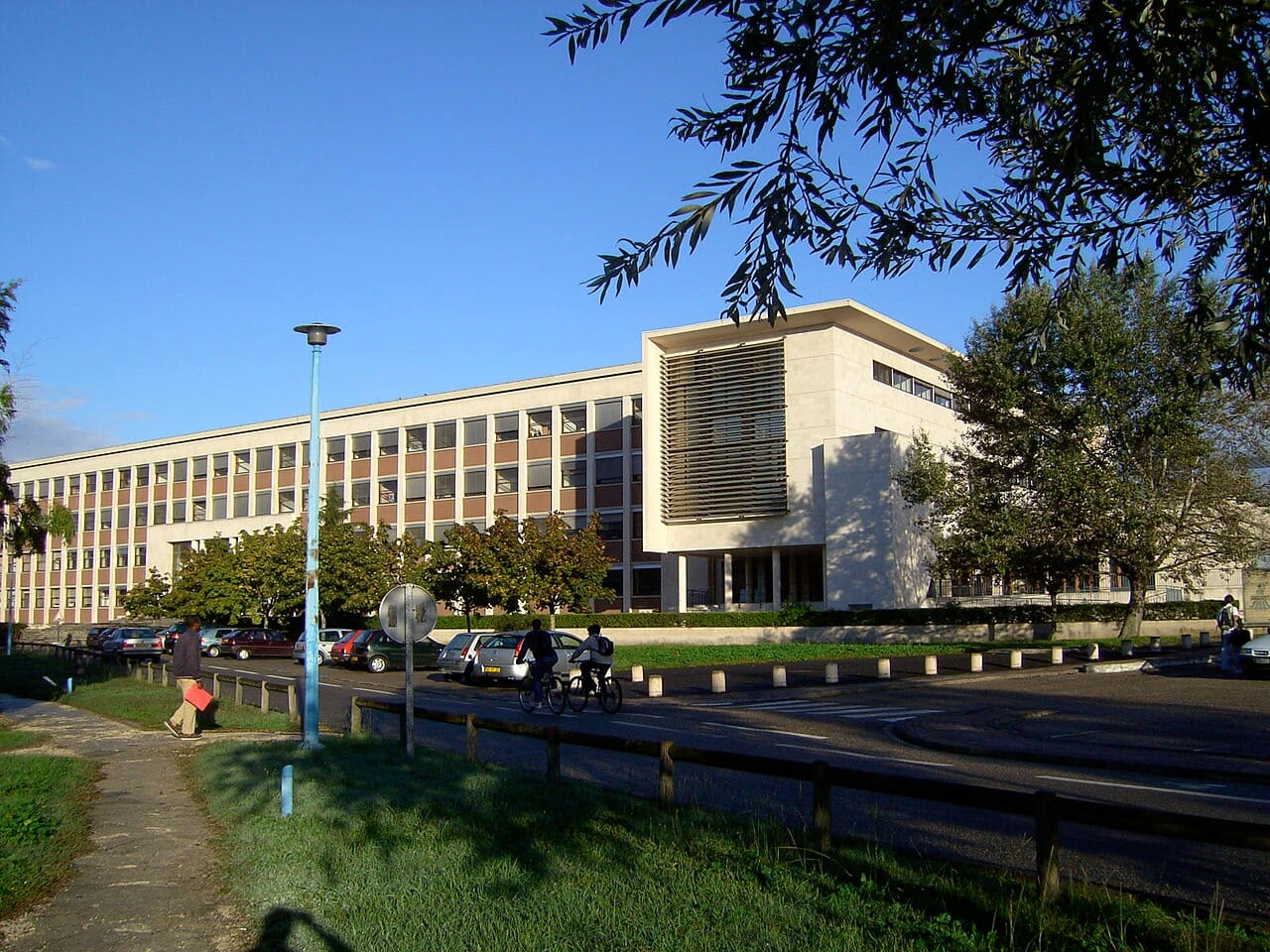 Bâtiment universitaire en façade claire sous un ciel bleu, avec des étudiants à pied et à vélo sur la route qui le longe