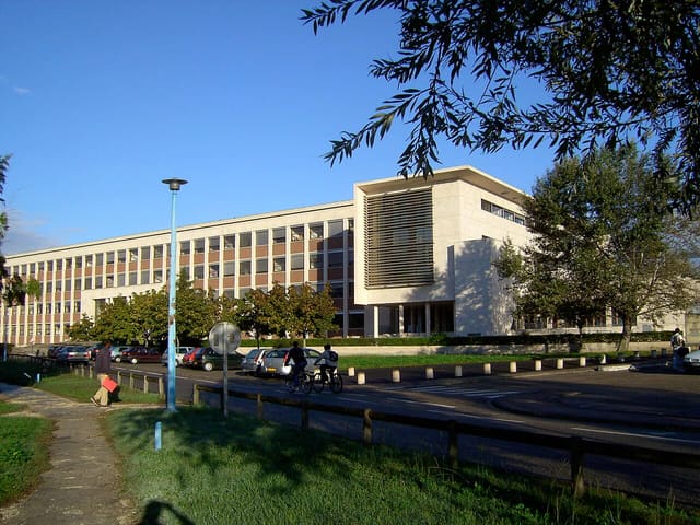 Bâtiment universitaire en façade claire sous un ciel bleu, avec des étudiants à pied et à vélo sur la route qui le longe
