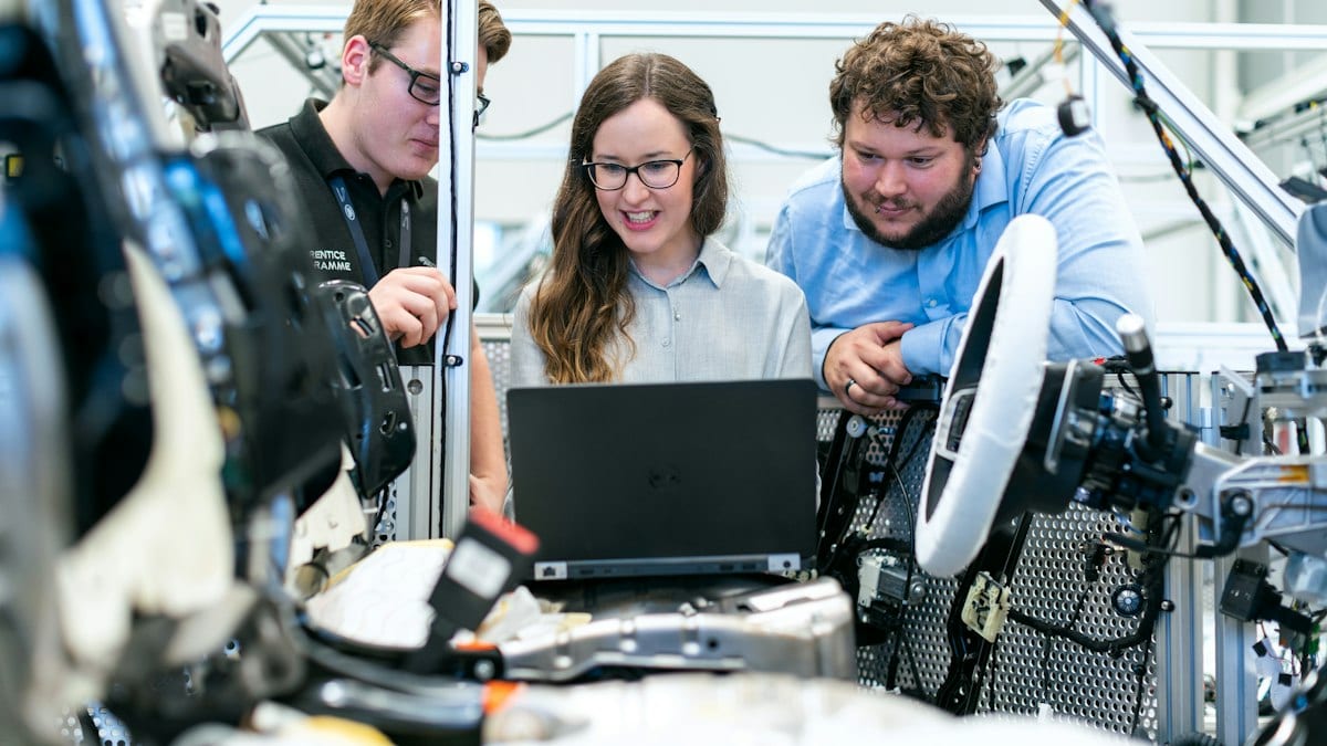 Trois jeunes ingénieurs penchés sur un ordinateur portable, entourés d'équipements mécaniques dans un atelier.