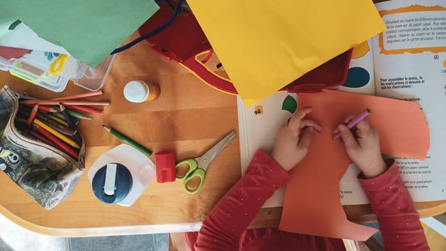 Vue de dessus d'un enfant qui découpe et colorie du papier cartonné sur une table couverte de crayons, de ciseaux et de colle.