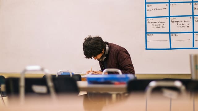 Un lycéen écrit à son bureau dans une salle de classe, devant un tableau blanc couvert de dates d'examens.