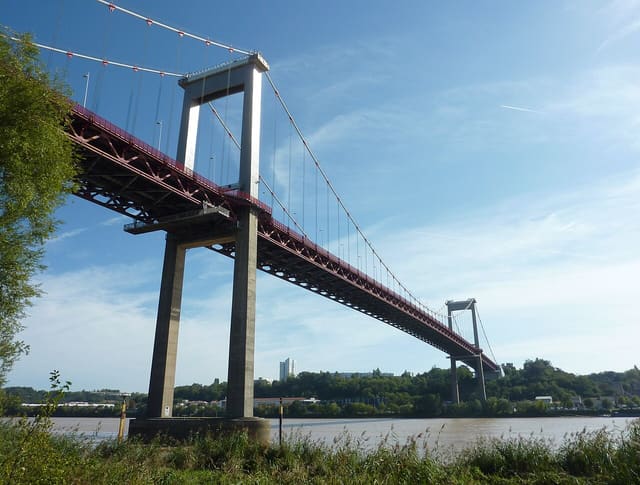 Le pont d'Aquitaine vu depuis la rive, avec son tablier rouge, ses pylônes en béton et ses câbles de suspension au-dessus de la Garonne.