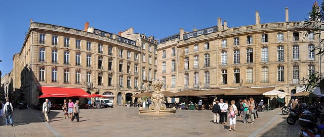 La place du Parlement à Bordeaux, avec sa fontaine centrale, ses façades de pierre blonde et ses terrasses de restaurants sous un ciel bleu.