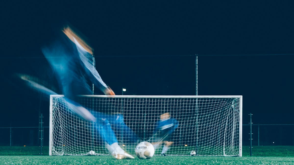 Un joueur frappe un penalty de nuit sous les projecteurs, le gardien plongeant vers le but, dans un flou de mouvement.