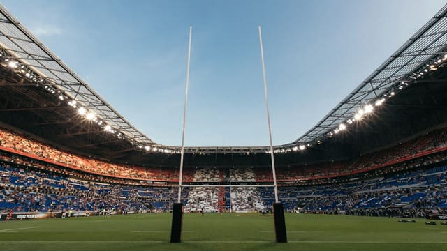 Vue depuis la pelouse d'un stade de rugby au crépuscule, avec les poteaux au premier plan et des tribunes bien garnies.