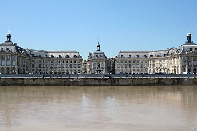 Façade du Palais de la Bourse et de la place de la Bourse à Bordeaux, vue depuis la Garonne, sous un ciel dégagé.
