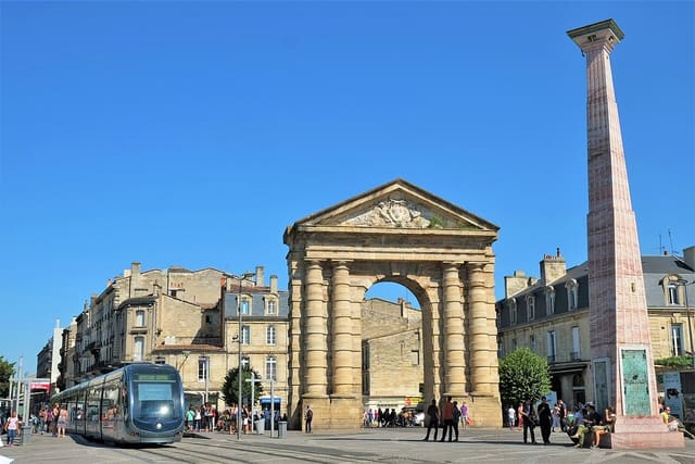 La porte d'Aquitaine et l'obélisque de la place de la Victoire à Bordeaux, un tramway et des passants sous un ciel bleu.