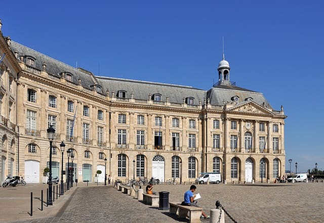 Façade du Palais de la Bourse de Bordeaux, bâtiment du XVIIIe siècle en pierre claire, vue depuis la place, sous un ciel bleu.