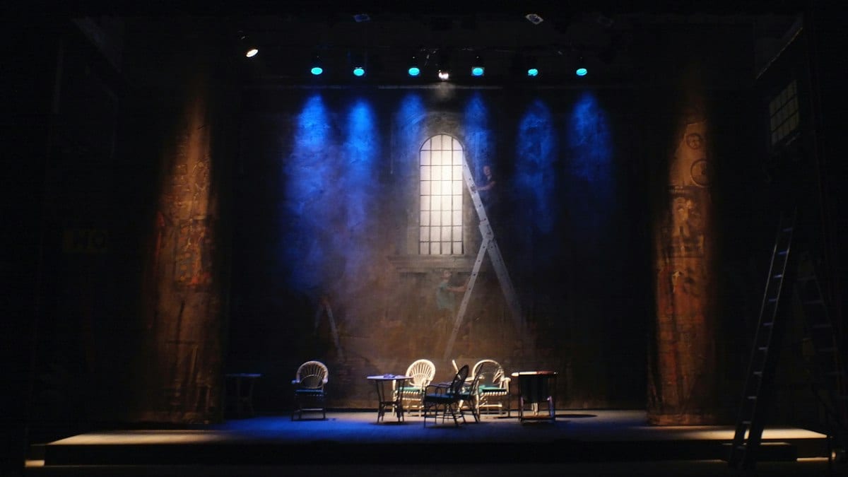 Scène de théâtre plongée dans une lumière bleue, avec quelques chaises et une échelle devant une grande fenêtre peinte