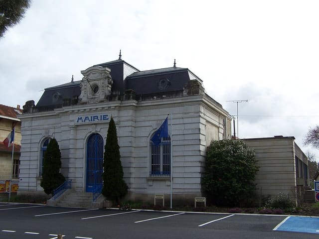 Façade d'une mairie en pierre blanche à toit d'ardoise, drapeaux français et européen devant l'entrée, parking au premier plan