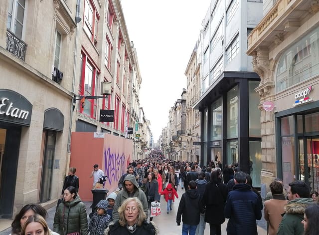 Piétons nombreux dans la rue Sainte-Catherine à Bordeaux, entre façades en pierre et enseignes de magasins.