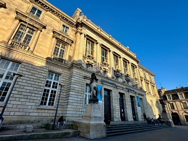 La façade historique de l'université de Bordeaux, place de la Victoire, avec des étudiants assis sur le parvis.