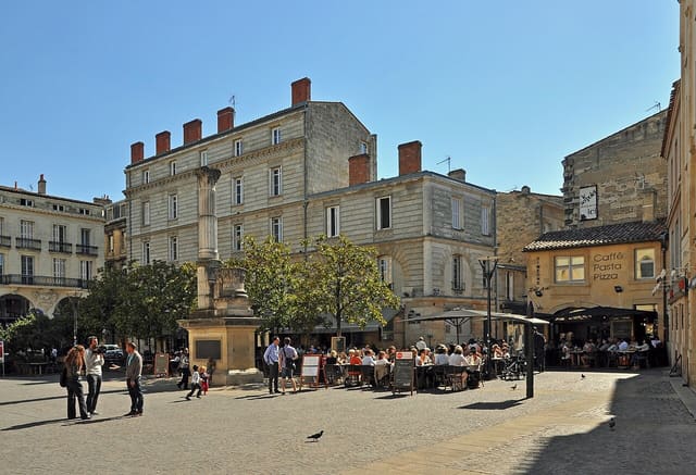 La place Camille-Jullian à Bordeaux, avec sa colonne, ses façades de pierre et les terrasses de café en plein soleil.